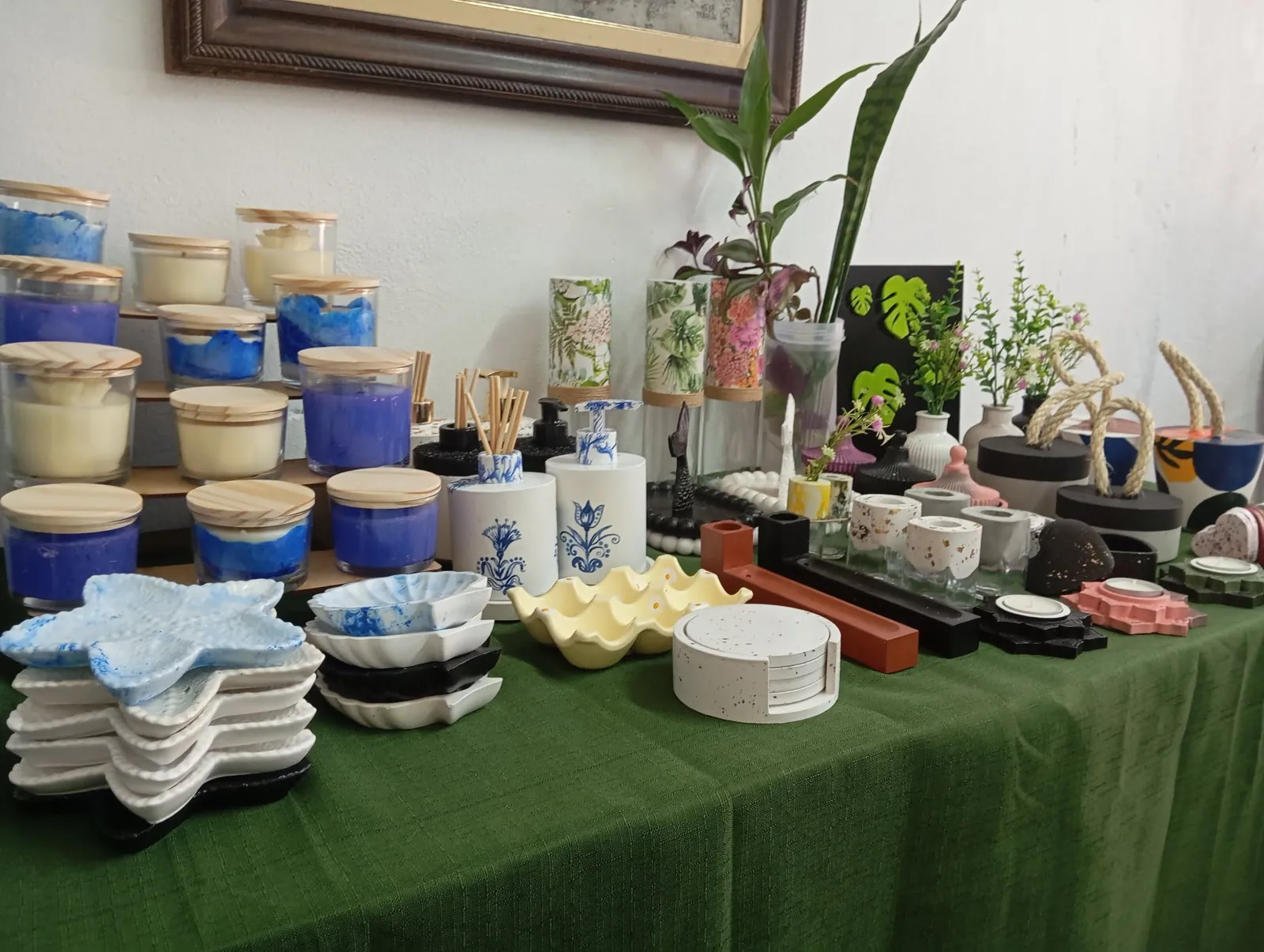 Mesa da Morada Ateliê numa feira: velas azuis, bandejas, porta-ovos, kits de lavabo, vasos e porta-copos sobre toalha verde