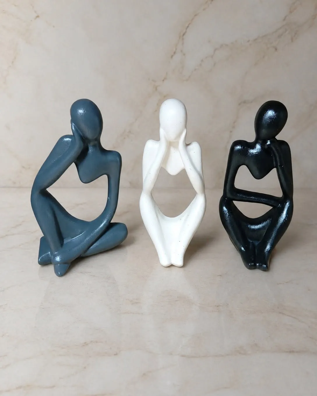 Três esculturas pensadoras sentadas, nas cores azul-petróleo, off-white e preto, sobre mármore